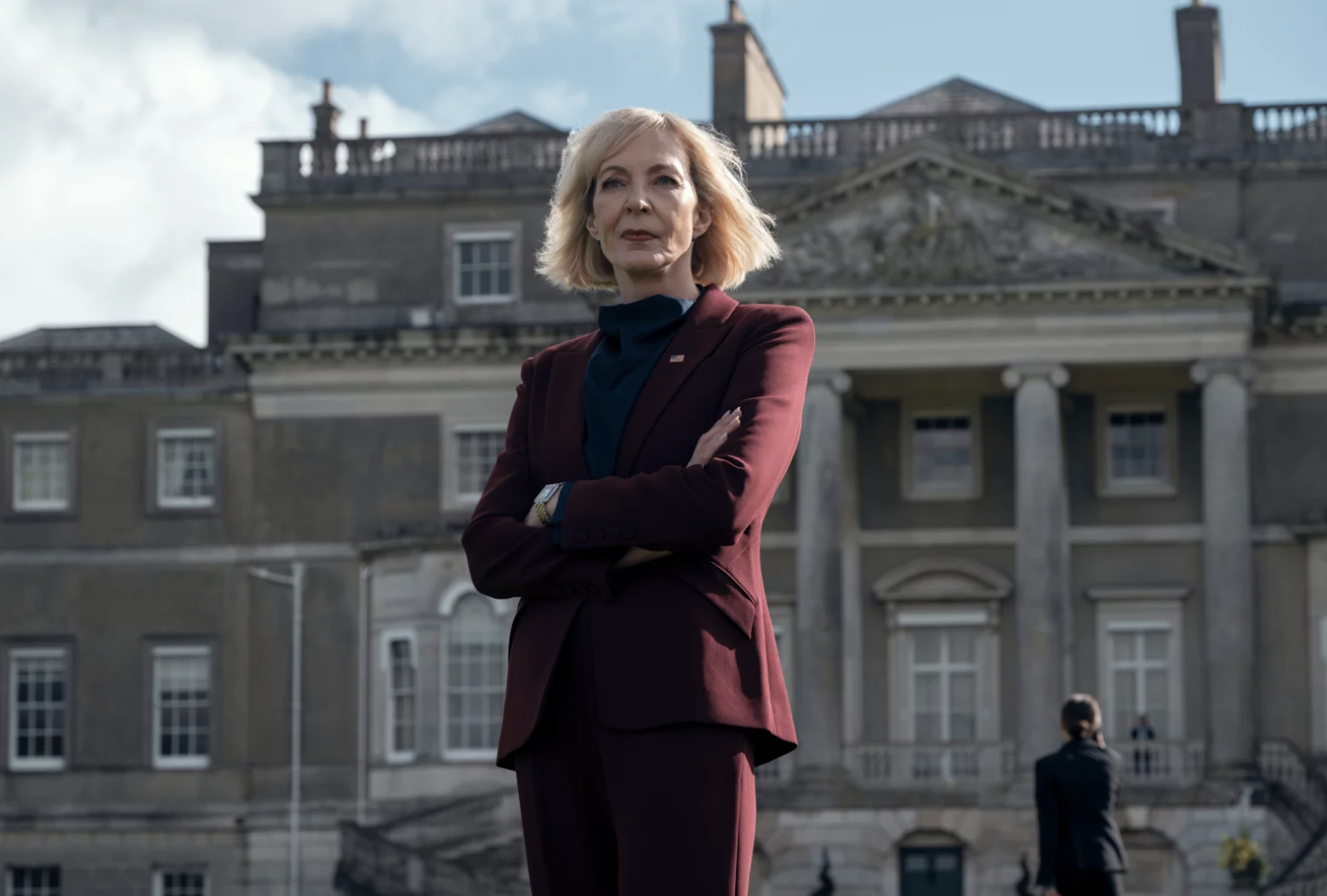 A middle aged woman with a blond bob stands, arms crossed, in front of a stately home. She is wearing a dark red pantsuit.