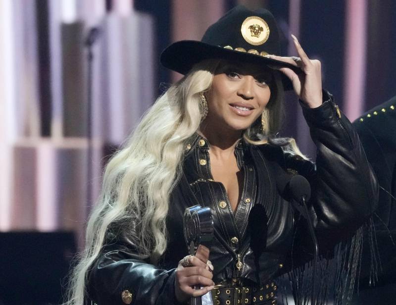 A Black woman with long white-blond hair tips her cowboy hat as she stands on stage holding a trophy. She is wearing all black.