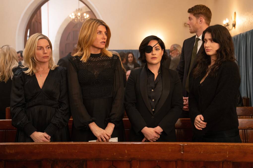 Four white women dressed in black, one wearing an eye patch, stand in a line in the pews of a church.