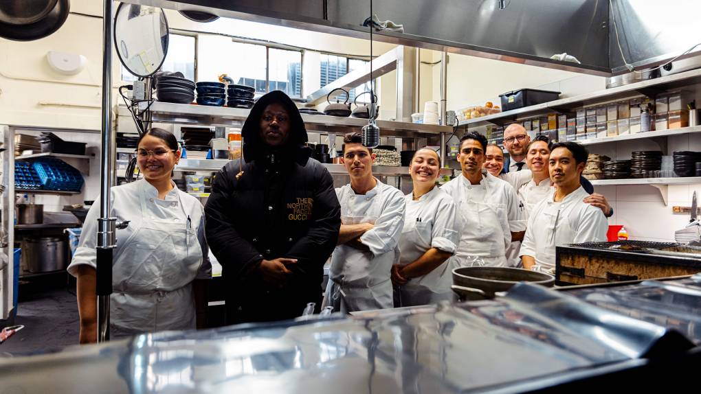 Rapper Blvck Svm, in a black puffy jacket, poses in the kitchen with the chefs of a fine dining restaurant, all wearing their white chef's jackets.