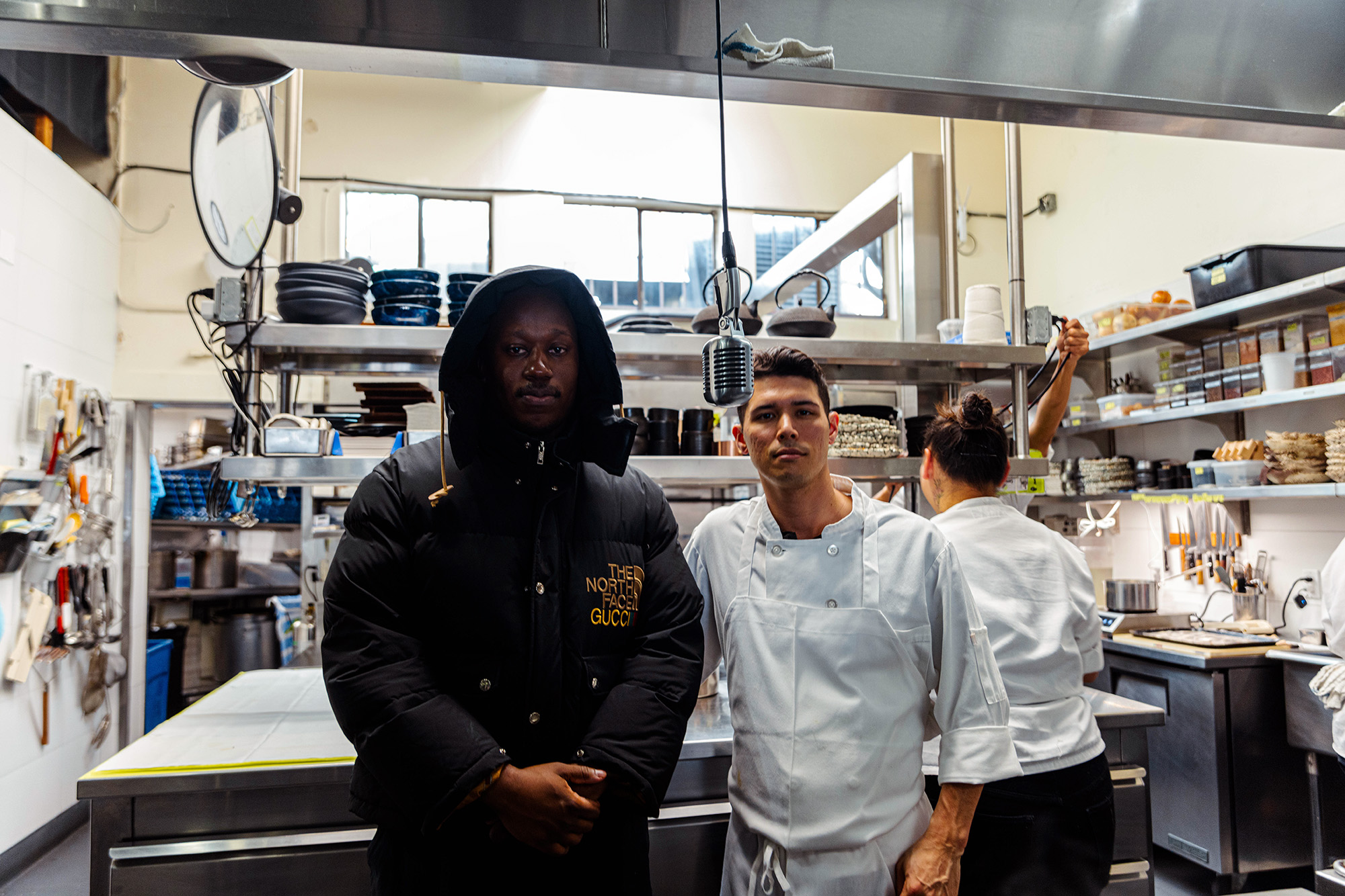 A rapper in a black puffy jacket poses with a chef in the kitchen of a fine dining restaurant.