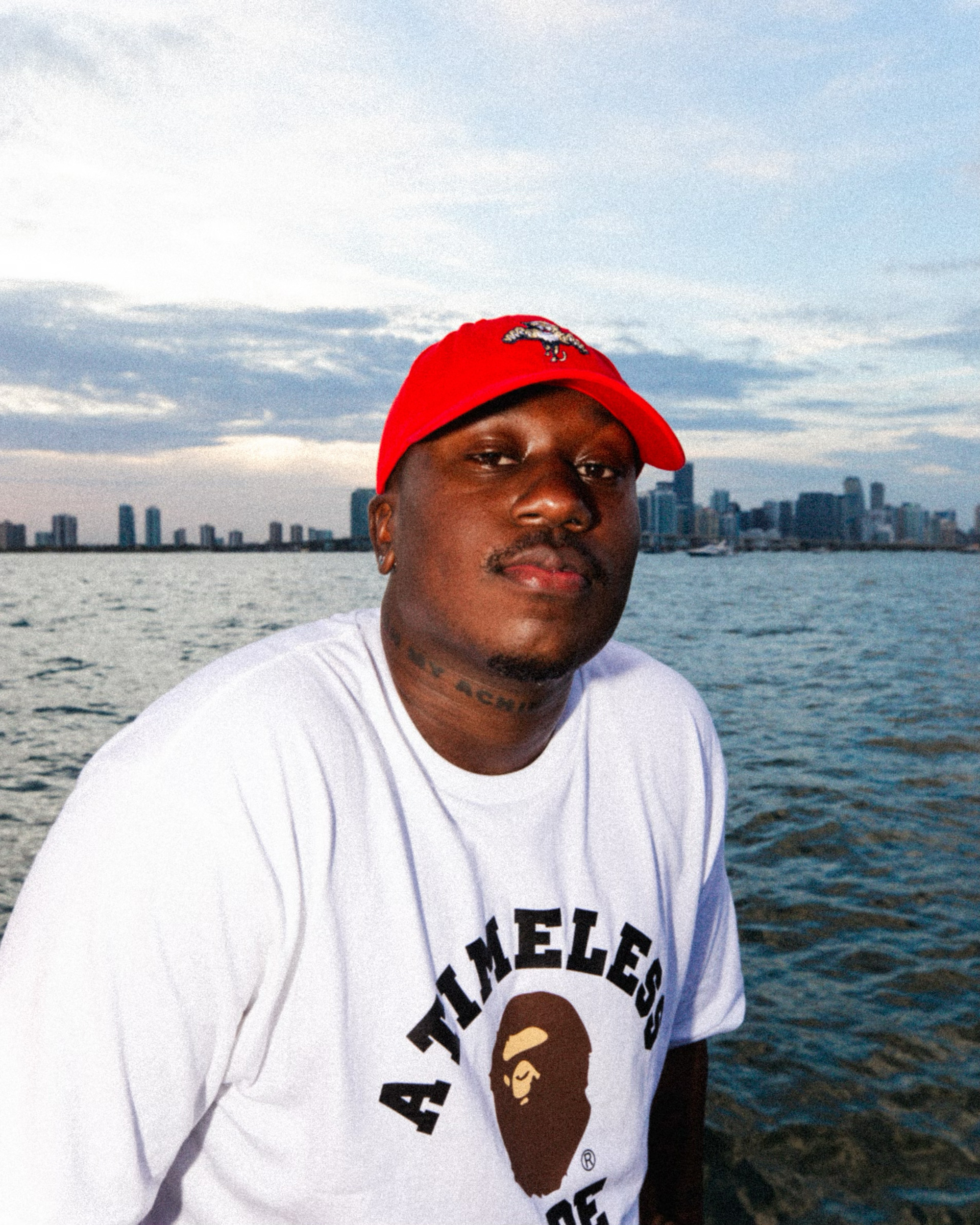 A man in a red baseball cap and white "A Timeless Ape" T-shirt poses in front of the Chicago waterfront.