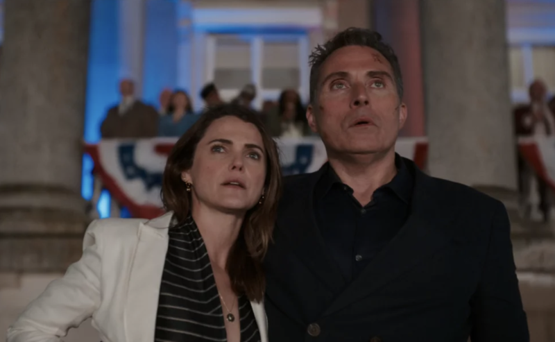 A white woman and white man stand in a grand room decorated patriotically, staring upwards and looking perplexed.