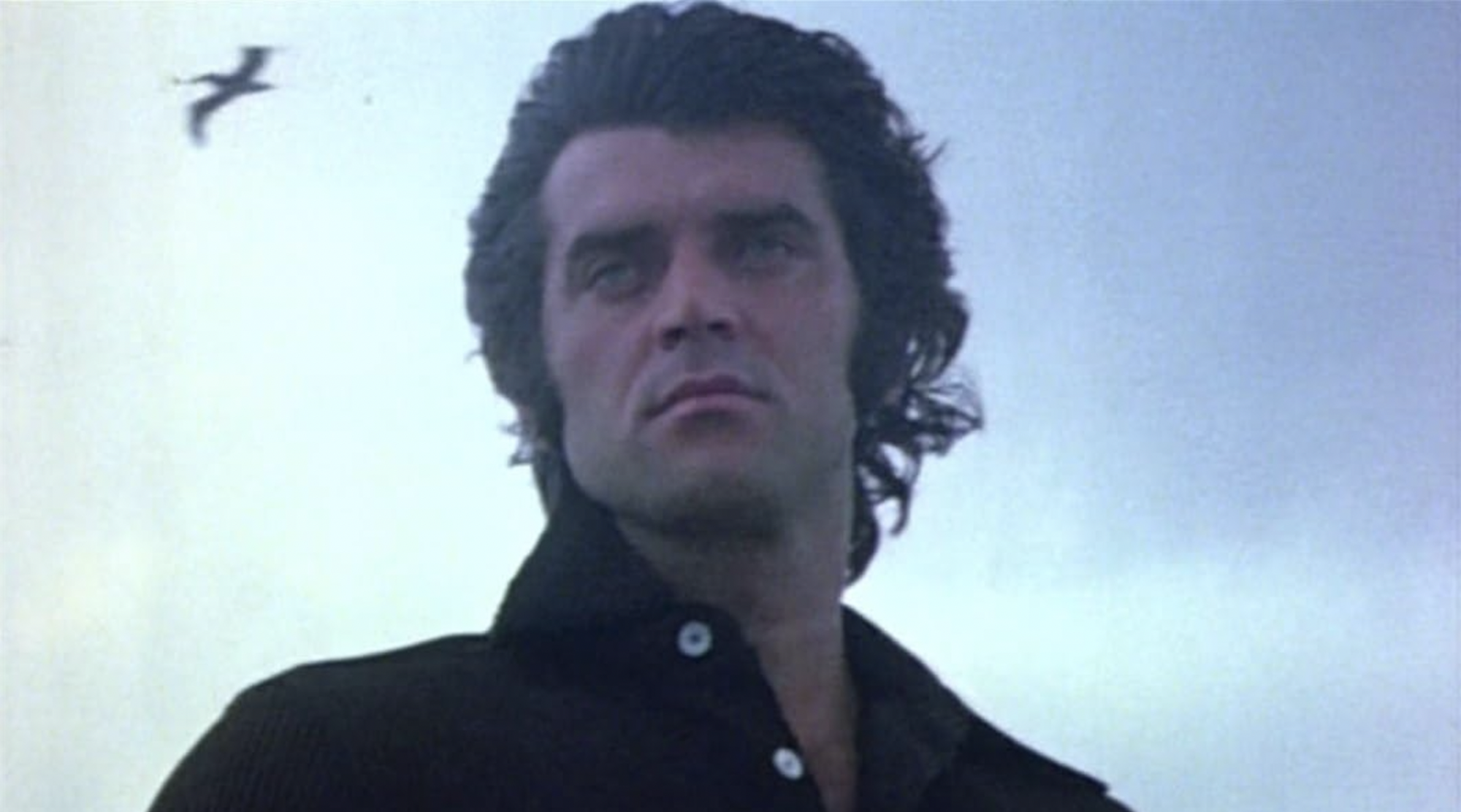 A brooding man wearing a black shirt stands, black hair blowing in the breeze.