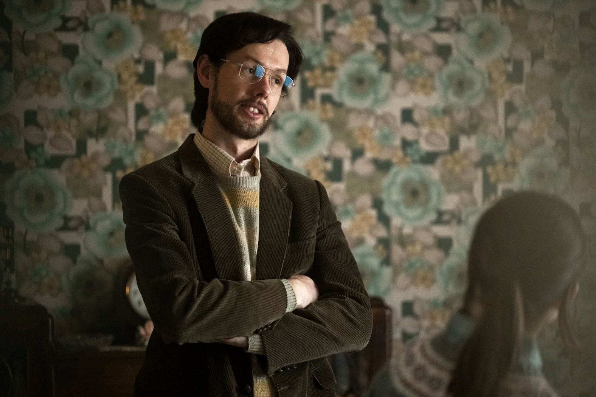 A white man with dark hair and beard stands with arms folded in front of 1970s-era wallpaper.