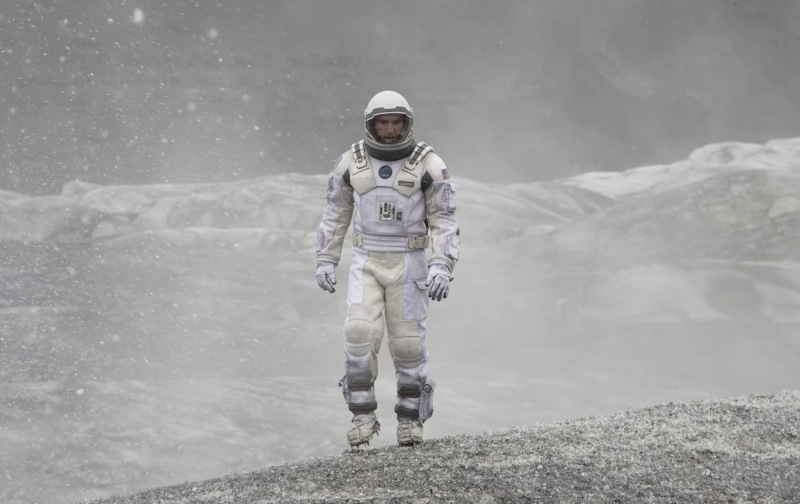 A man in a white space suit and helmet walks across mounds of grey and white gravel.