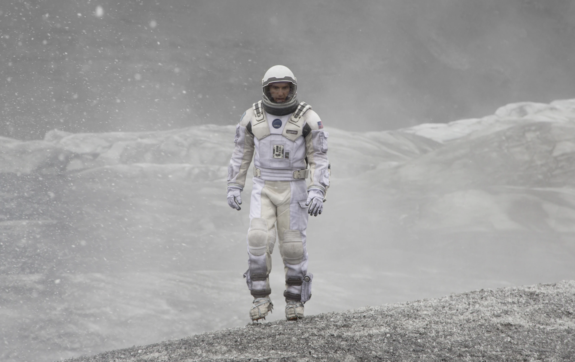 A man in a white space suit and helmet walks across mounds of grey and white gravel.