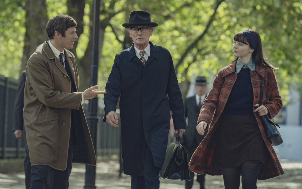 Two men in overcoats and a woman in a dark day dress walk in a line, engaged in conversation, under a canopy of trees in a city street.