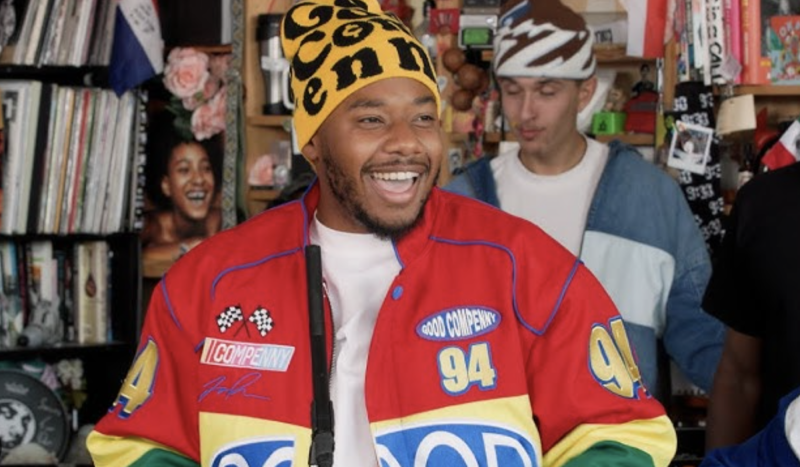 A smiling Black man wearing a red racing jacket and yellow and black beanie. Behind him are tightly packed book shelves and another man.