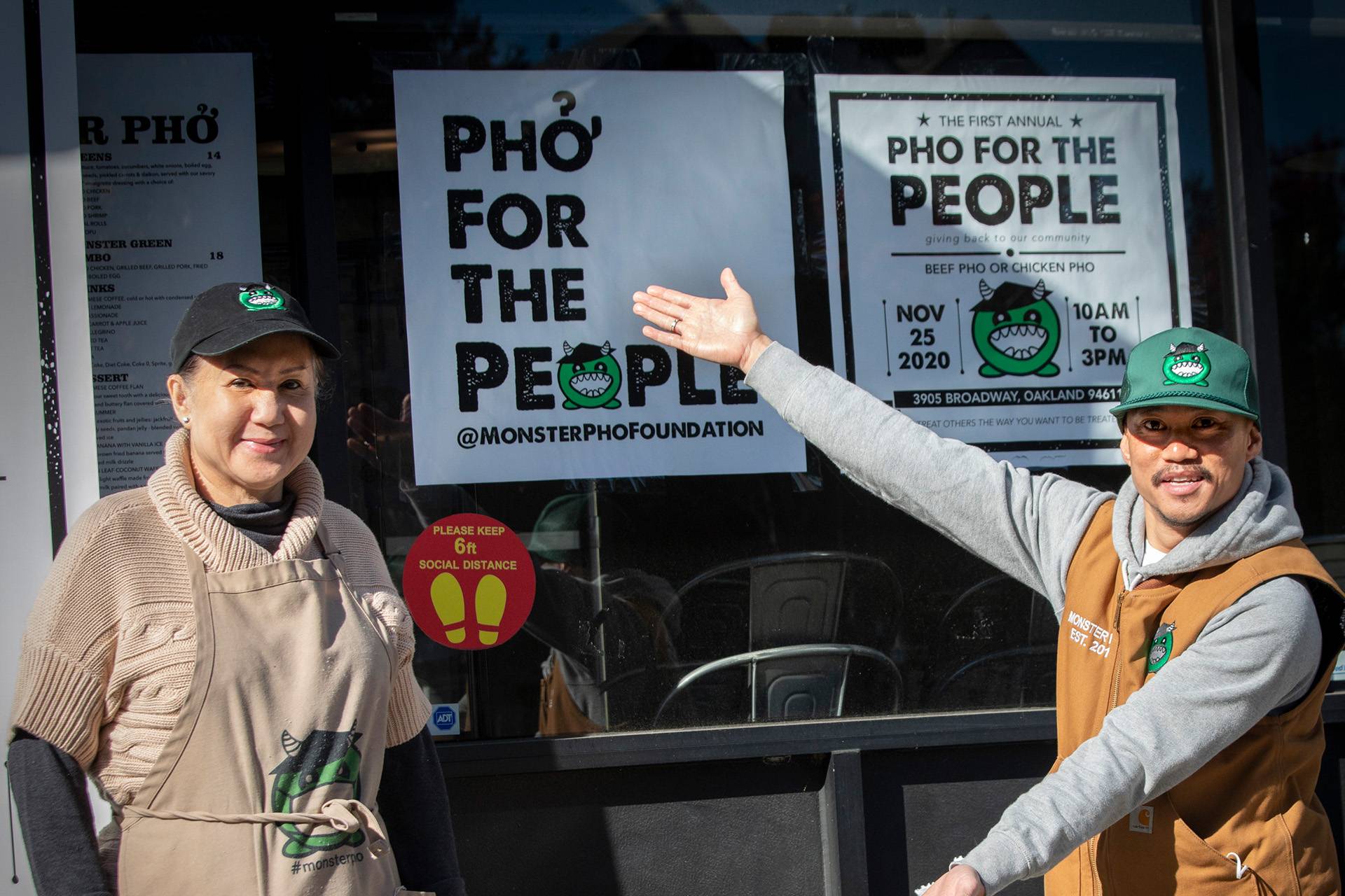 A man gestures toward a sign that reads, "Pho for the People." An older woman next to him smiles for the camera.