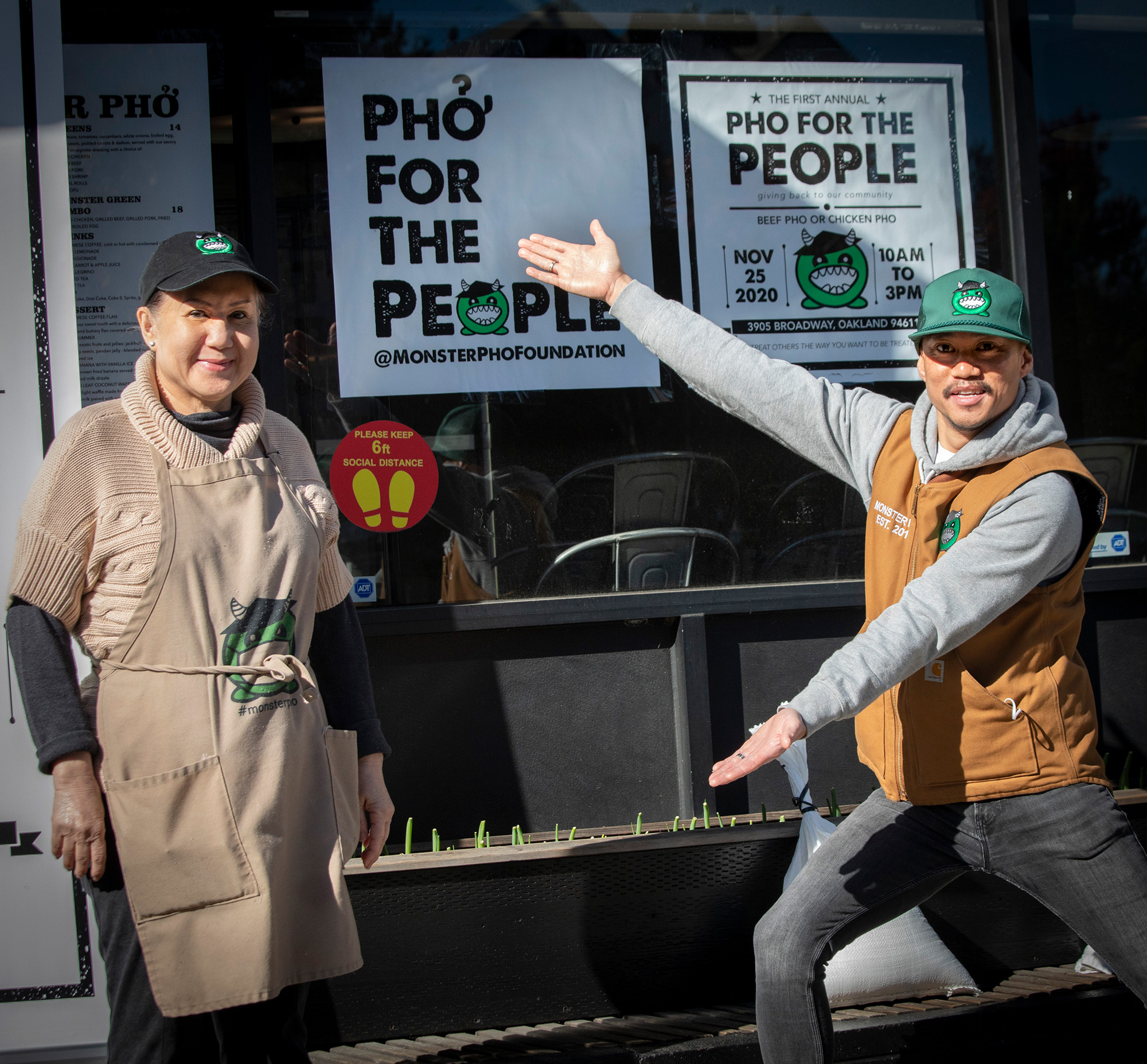 A man gestures toward a sign that reads, "Pho for the People." An older woman next to him smiles for the camera.