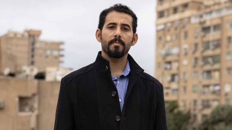 Man in dark jacket pictured in front of tall brown buildings