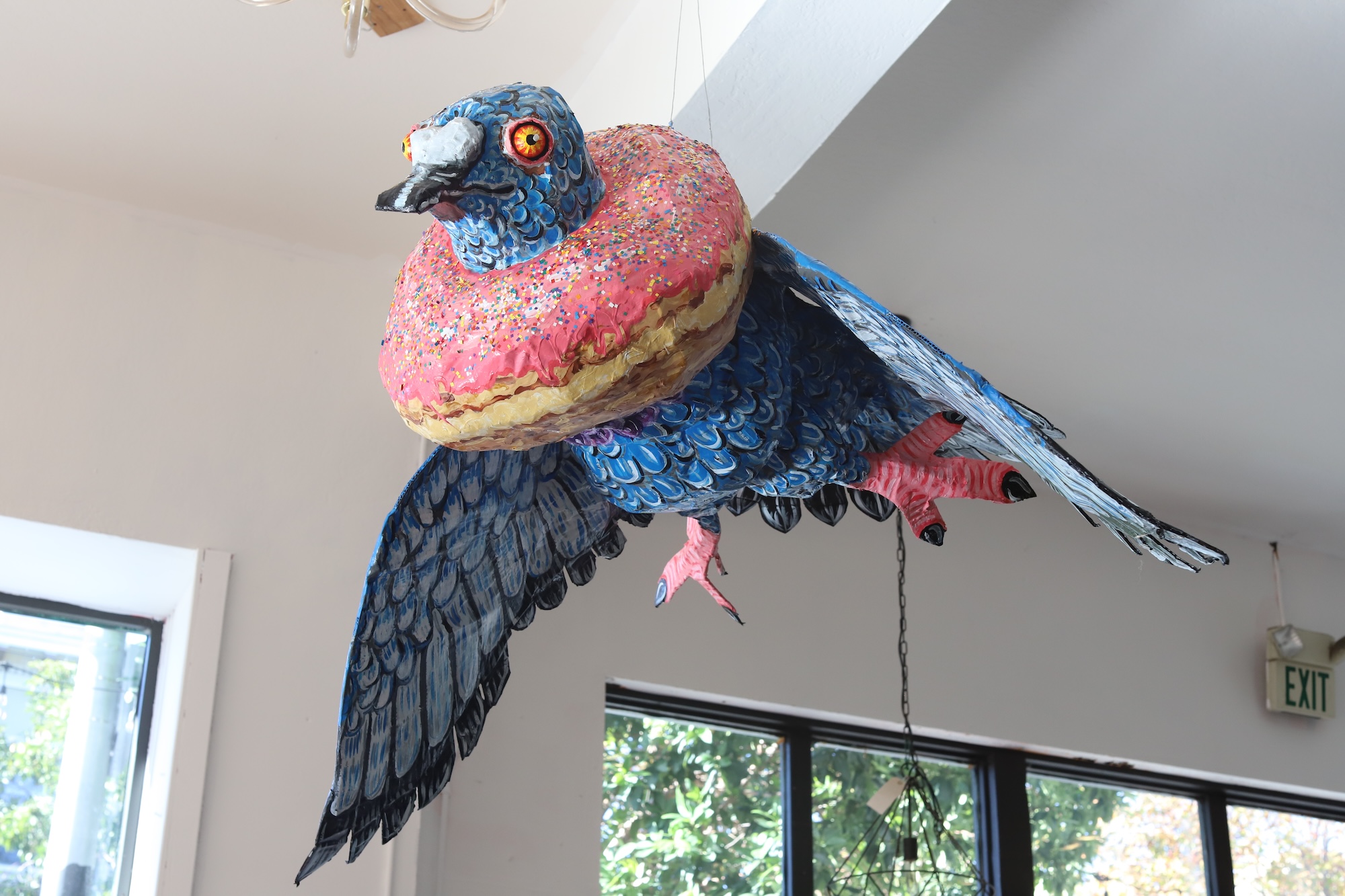 A giant paper mache blue pigeon mid-flight with a pink donut around its neck.