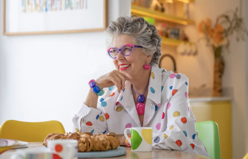 A senior woman sits at a kitchen table wearing a polka-dotted white jacket and colorful accessories. She is smiling. Mugs and a plate of croissants sit before her on the table.