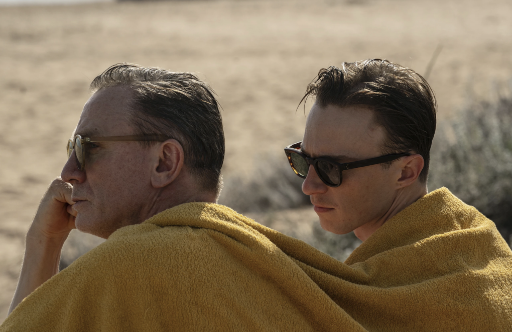 Two men sit under a yellow blanket on a beach.