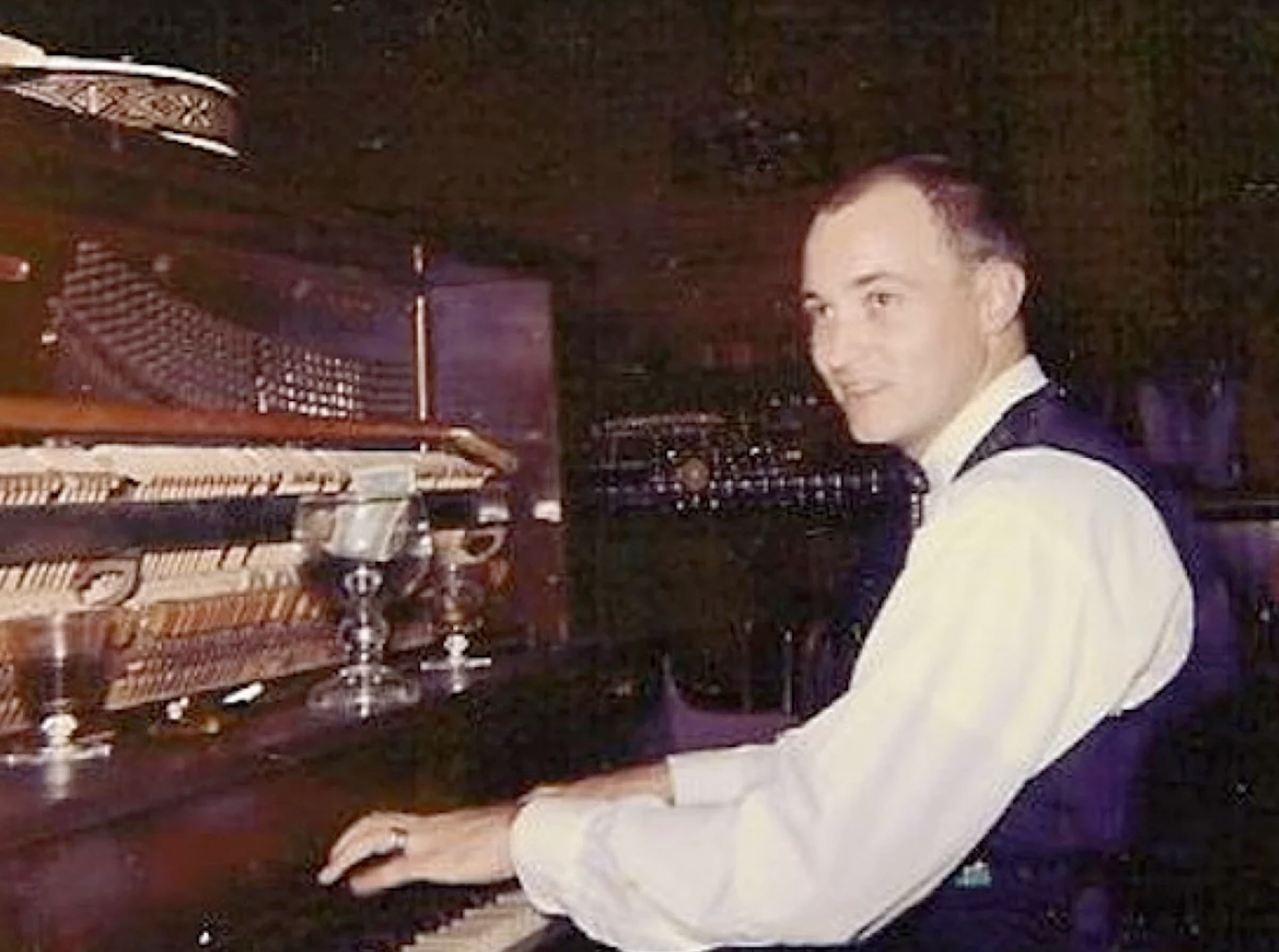 A young man in a shirt and vest plays a piano inside a bar.