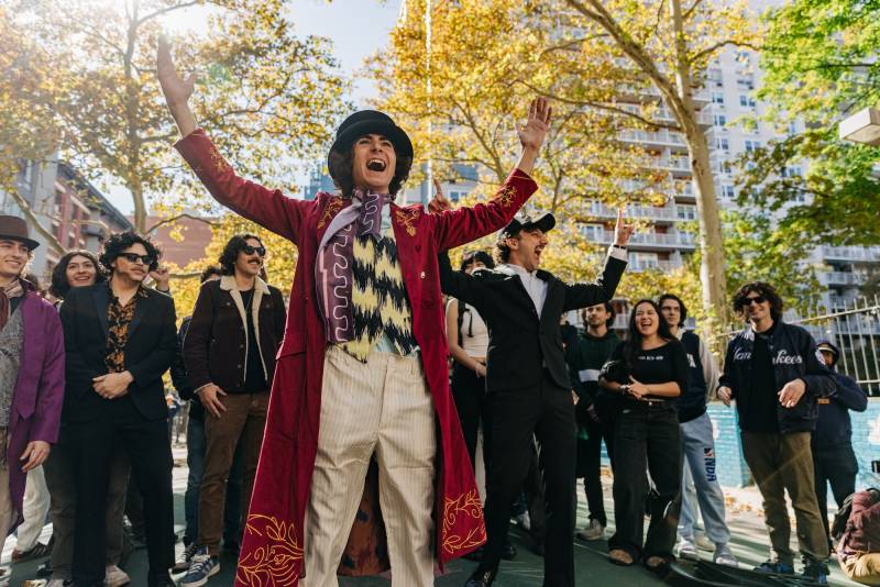 A young man dressed as Willy Wonka raises his arms and cheers, surrounded by crowds in a park.