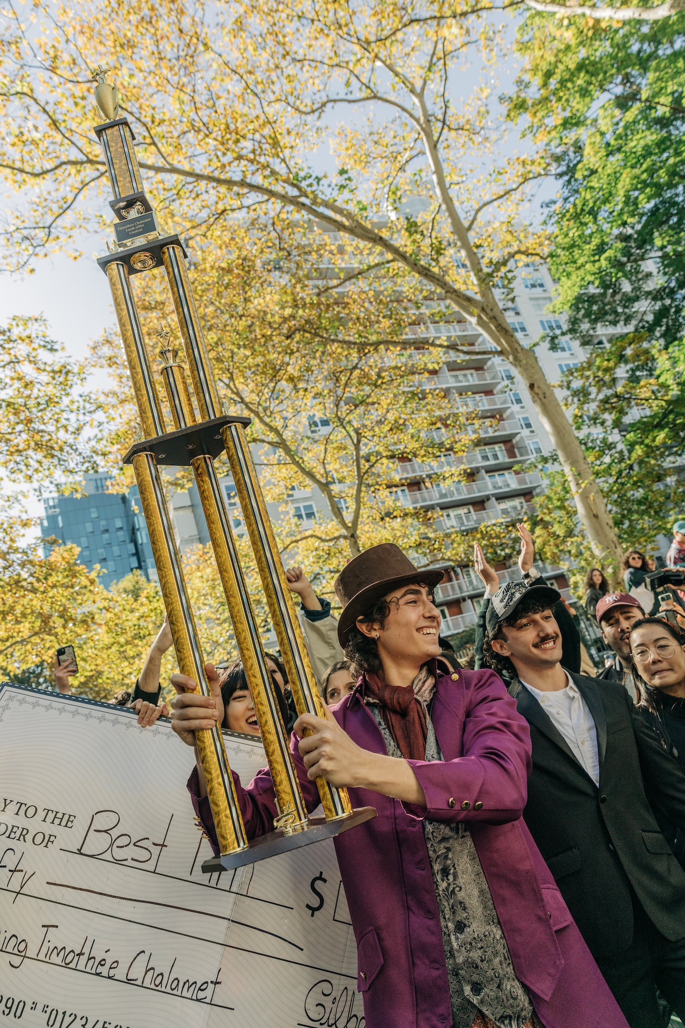 A young man holds up a 4ft tropny in a park while dressed like Willy Wonka.