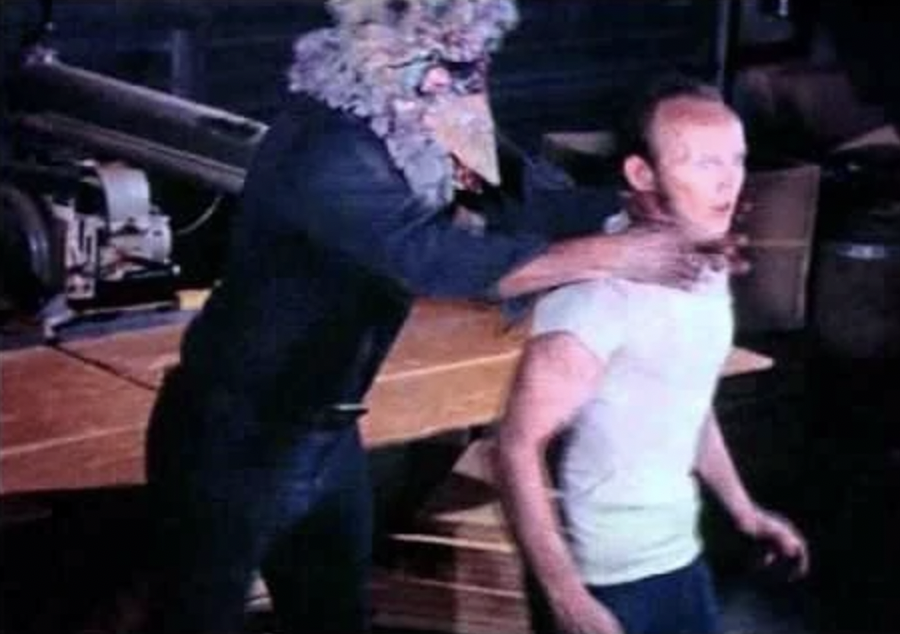 A man in a white t-shirt and jeans stands stunned while a man with a turkey head grips him by the neck.