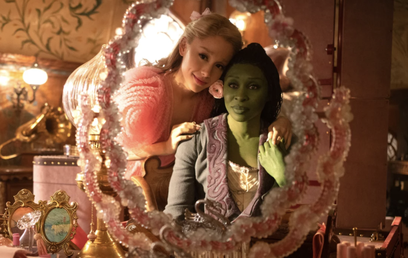 A white blond woman dressed in pink rests her hand on the shoulder of a green woman dressed in black sitting at a vanity mirror.