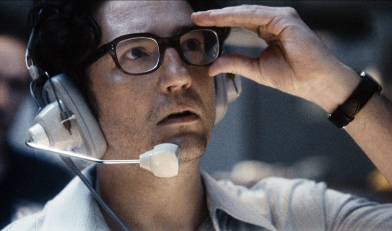 A man wearing an old-fashioned headset and microphone stares upwards, looking very concerned, while clutching the side of his black-rimmed glasses.