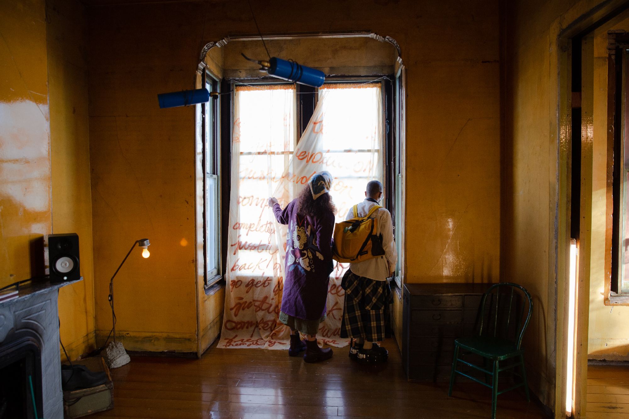 two figures hold gauzy curtain with writing aside to look out window from living space