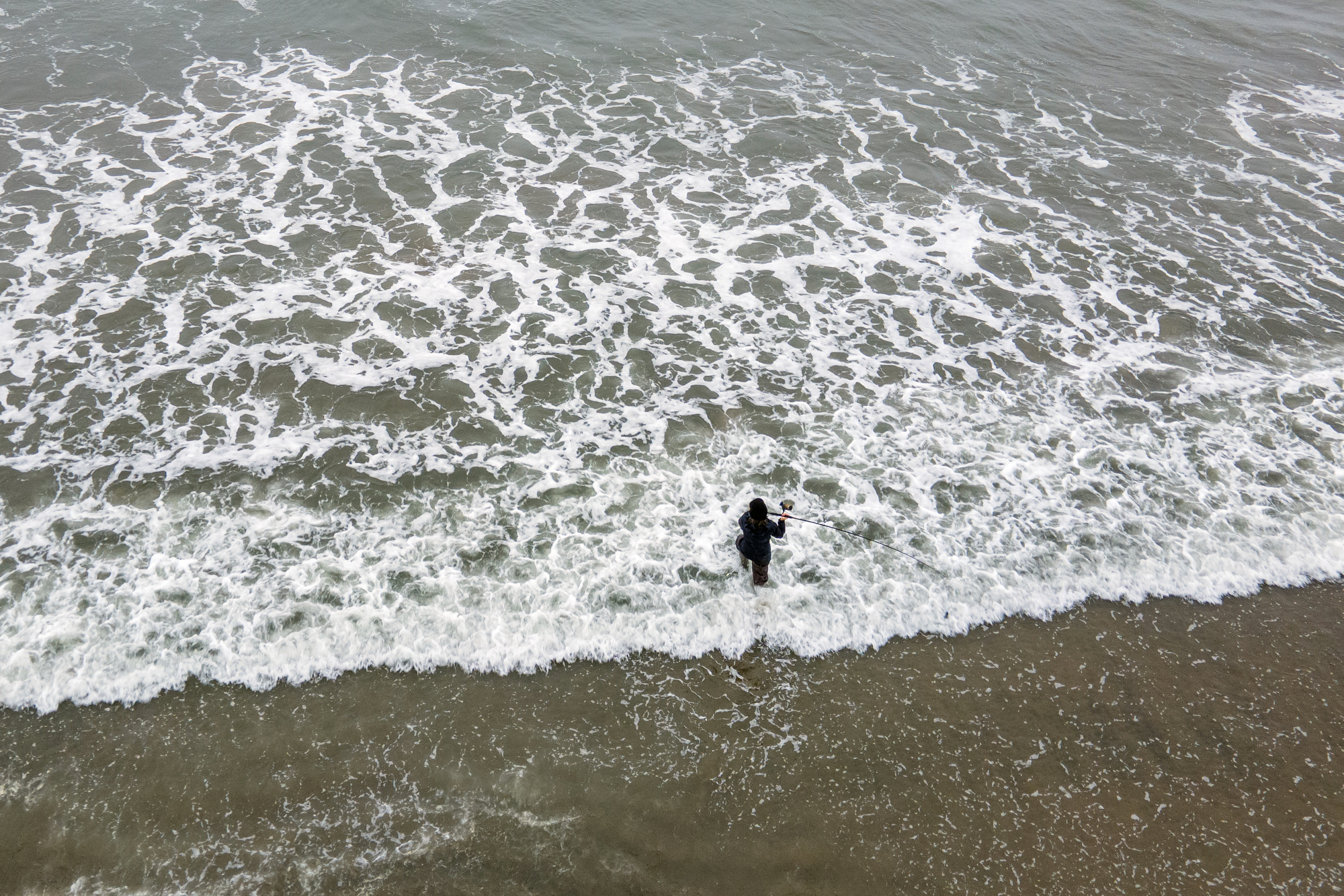 Overhead drone photo of a fisherman wading into the choppy waters on the edge of the beach.