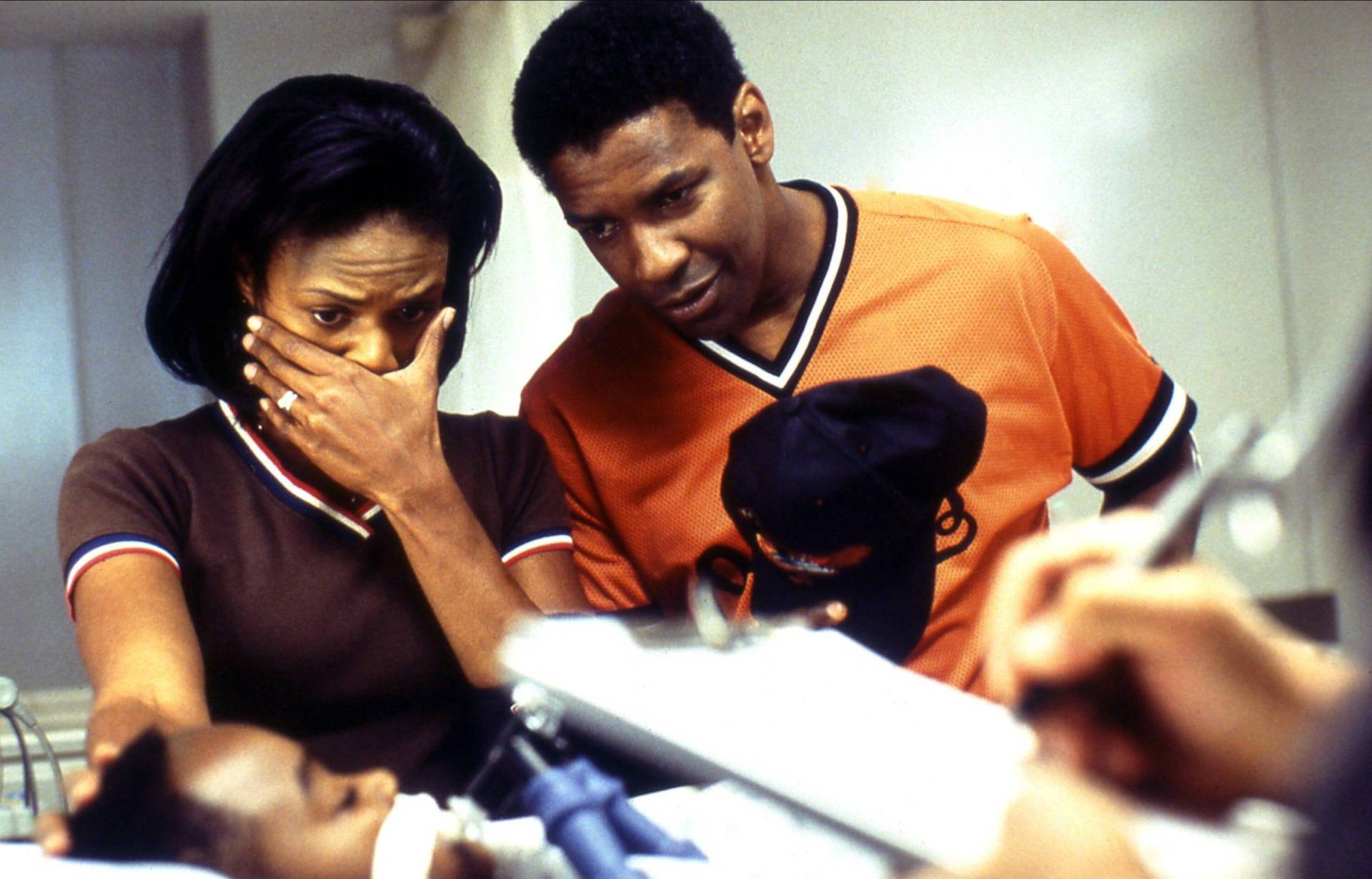 An attractive Black couple stand over a sleeping Black child in a hospital bed. The woman holds one hand over her face emotionally.