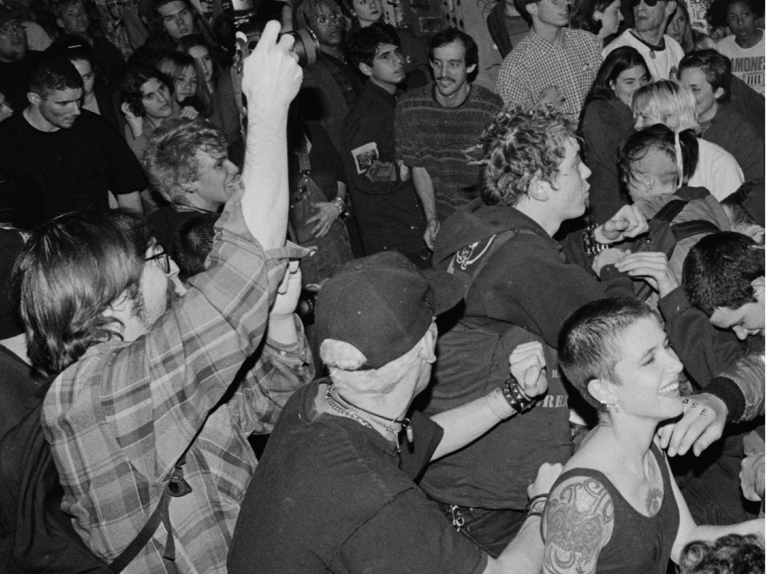 A black and white crowd shot at a punk show. Photographer Murray Bowles, a bearded white man in a flannel shirt, is featured mid-frame with his camera raised above his head.