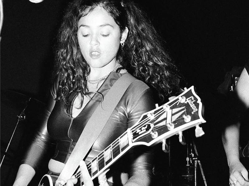 A young white girl with long brown hair plays a Gibson guitar