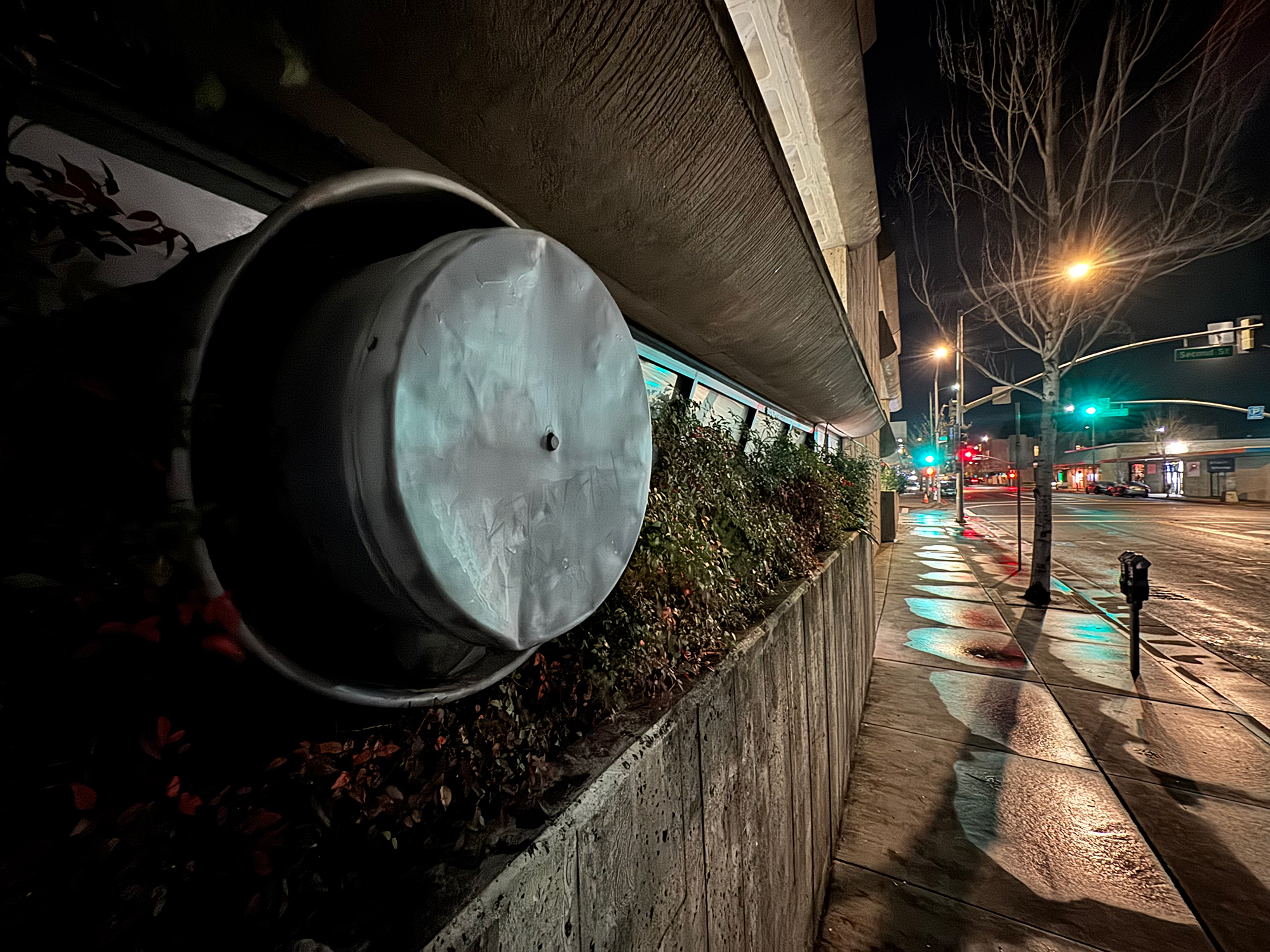A circular metal casing sticks out of a concrete wall, with foliage growing around it, and the nighttime glow of traffic lights in the background