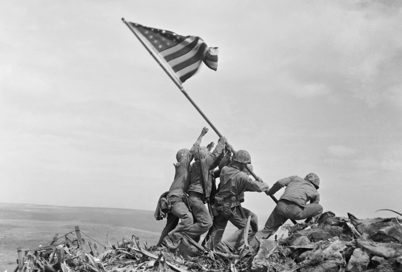 Four men in military uniforms, their faces obscured, struggle atop piles of shrapnel together to raise an American flag.