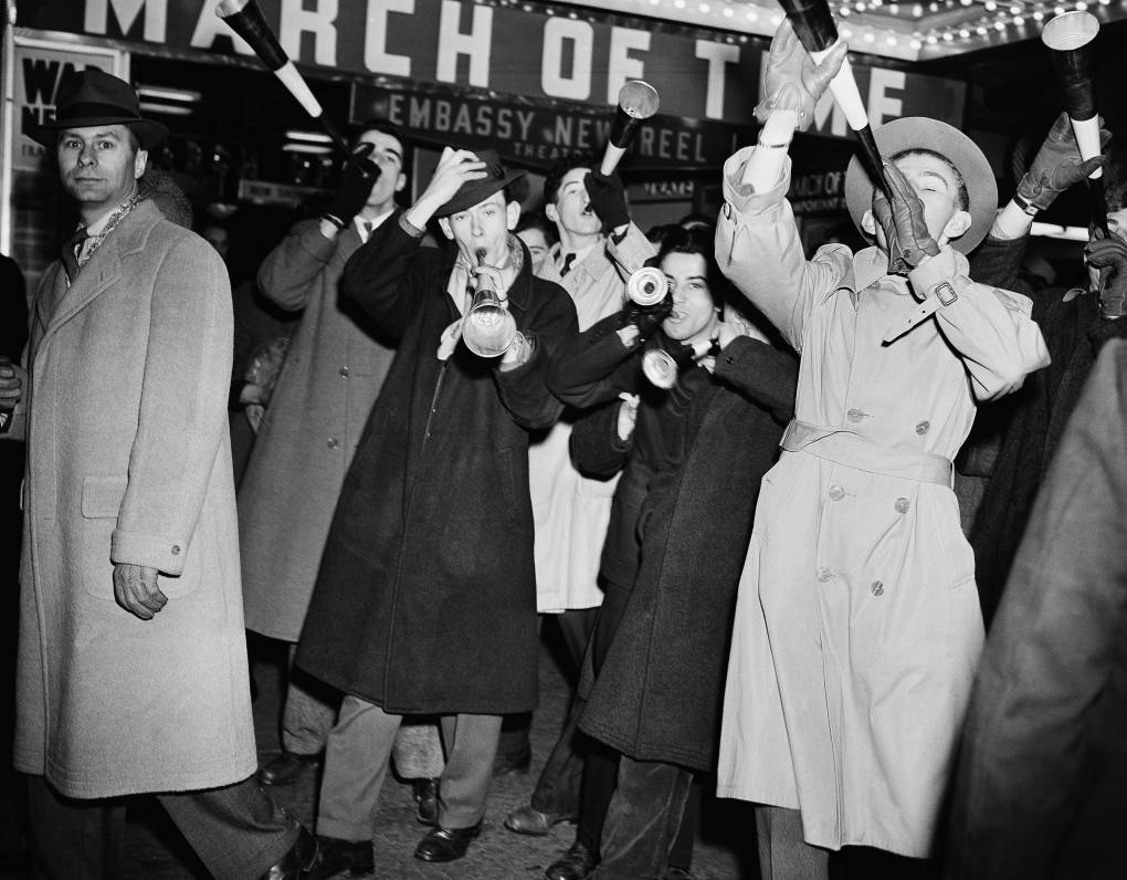 Men in 1940s attire — raincoats and hats — stand close together and blow novelty festive trumpets.