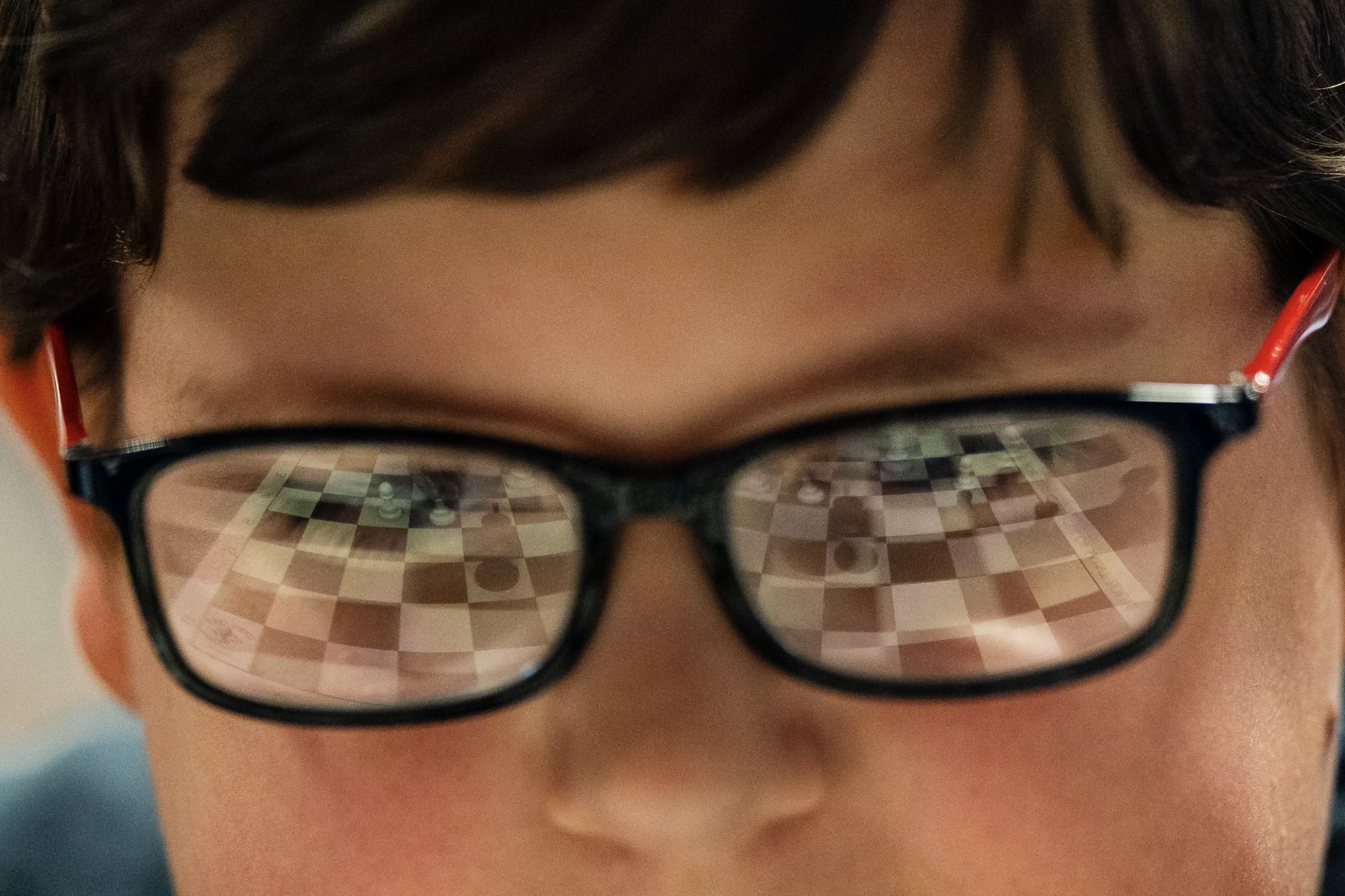 A close up image of a small boy wearing black rimmed glasses. A chess game is reflected in the lenses.