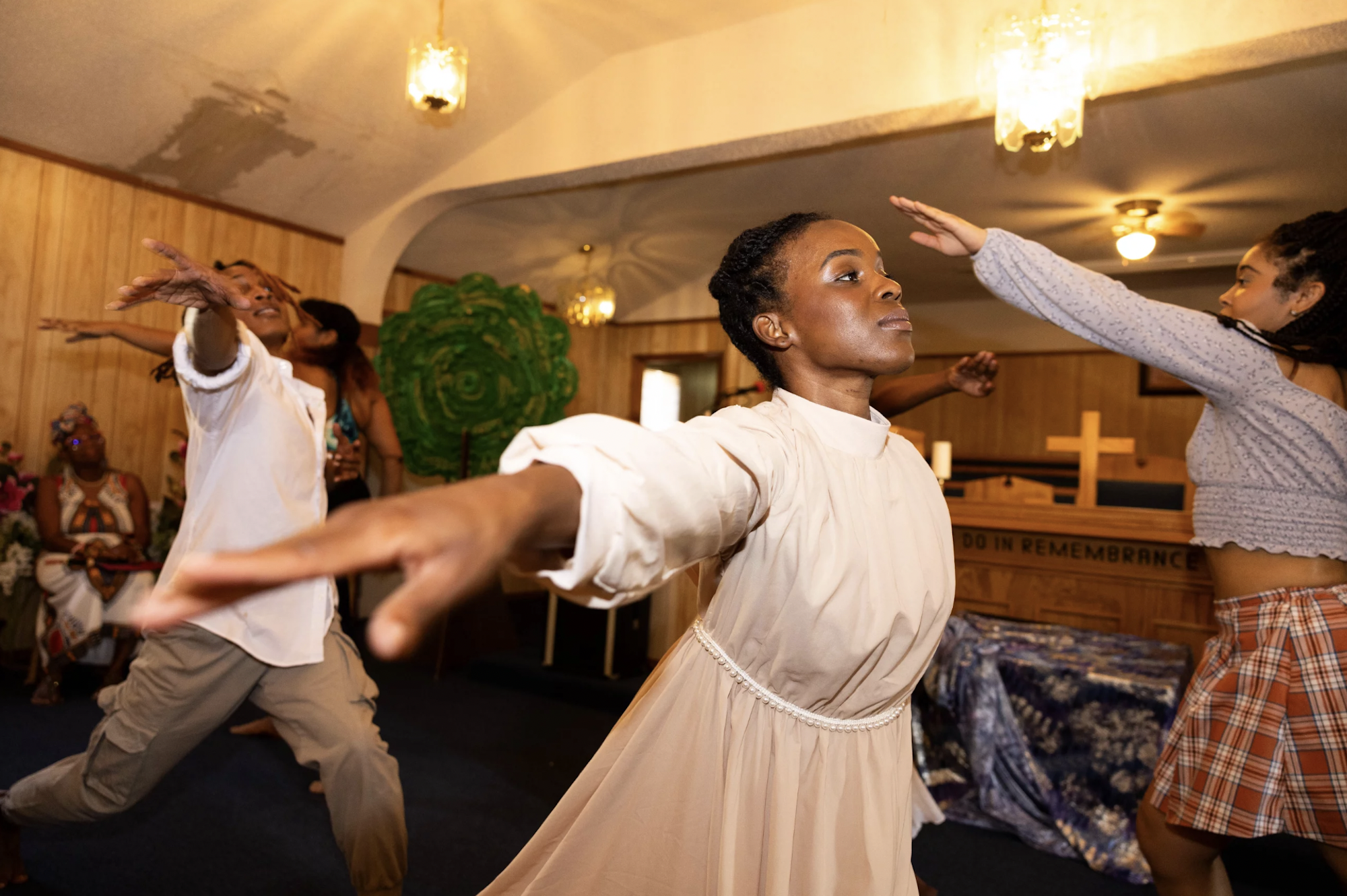 A Black woman wearing an old-fashioned white smock stretches out her arms as she performs with two other dancers at an indoor venue.