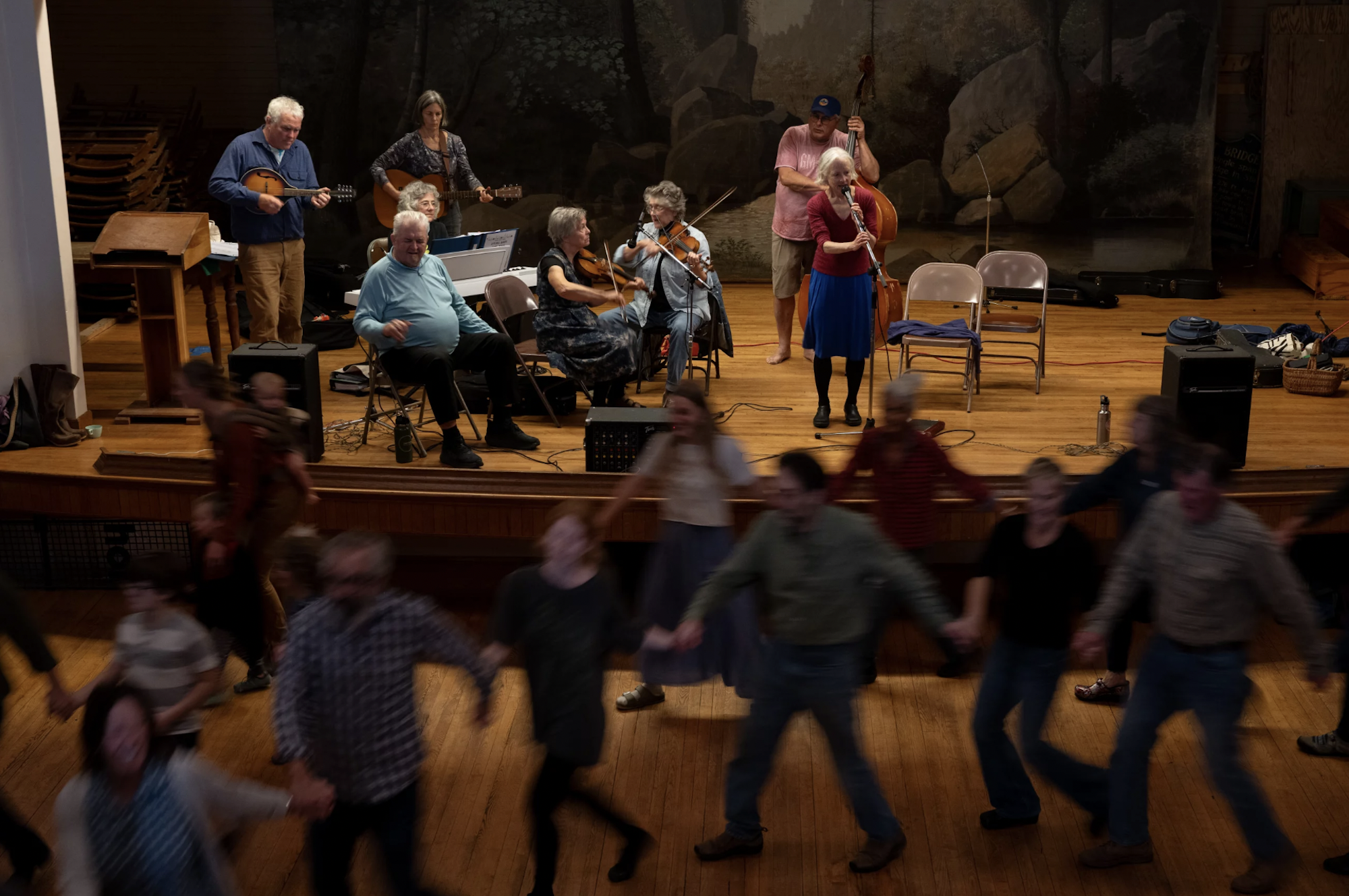 Dancers hold hands and move in a circle as a band plays on a stage nearby.