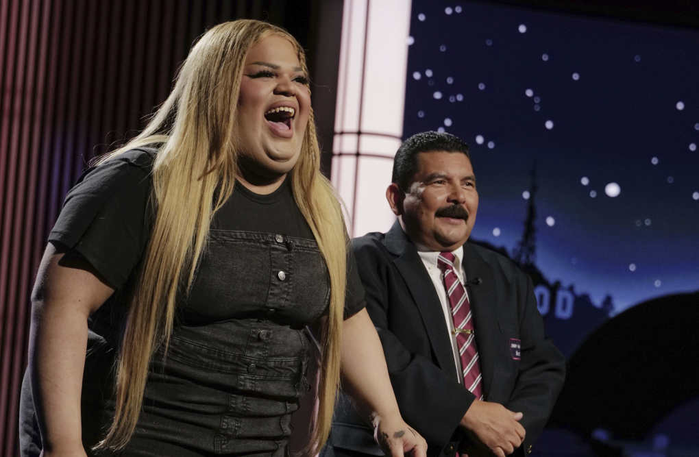 A rotund Black woman with long blonde hair in a tight black denim dress laughs uproariously on a TV set. A shorter Latino man wearing a tie and blazer stands near her.