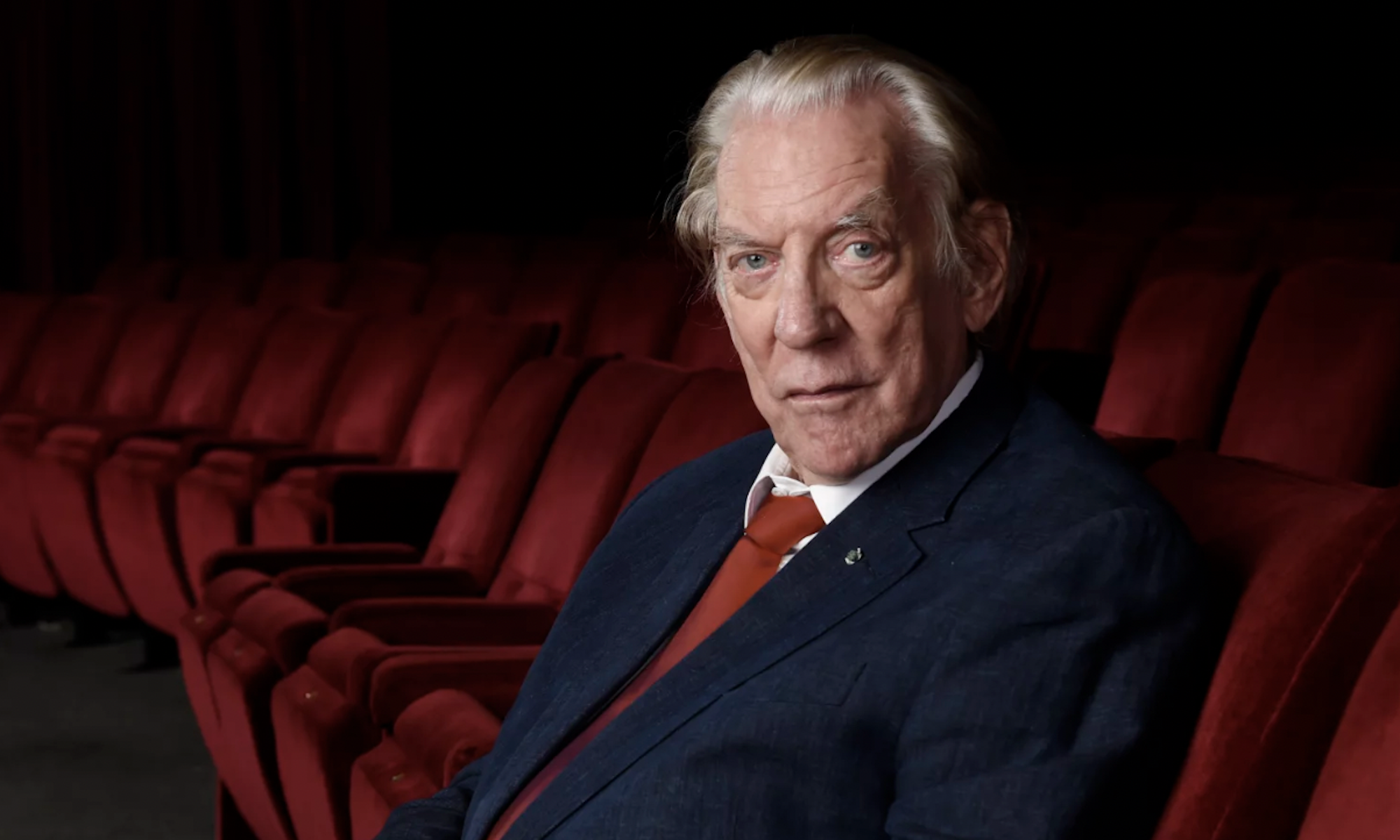 A senior man with white hair sits in a row of red theater seats. He is wearing a blue suit and red tie.
