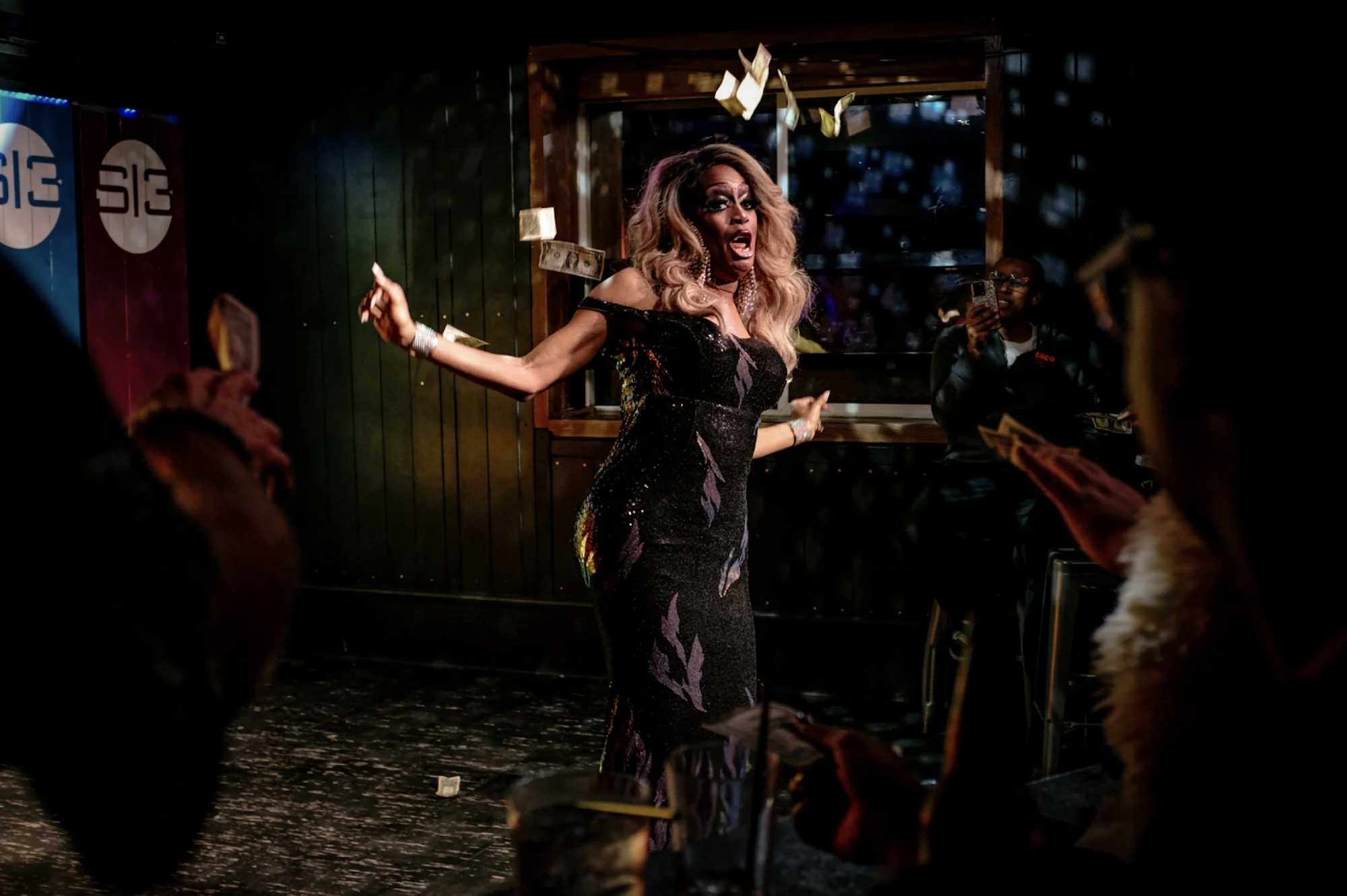 A Black drag queen performs and gestures while wearing a tight-fitting black gown inside a nightclub.