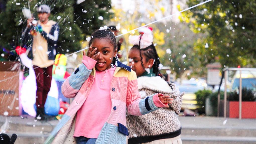 Children in winter coats enjoy artificial snowfall at an outdoor festival.