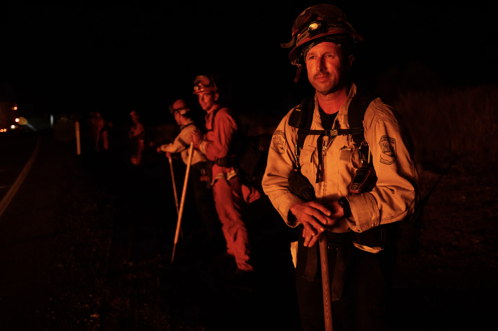 A line of firefighters stand in the darkness, their faces lit up by unseen flames in the distance.