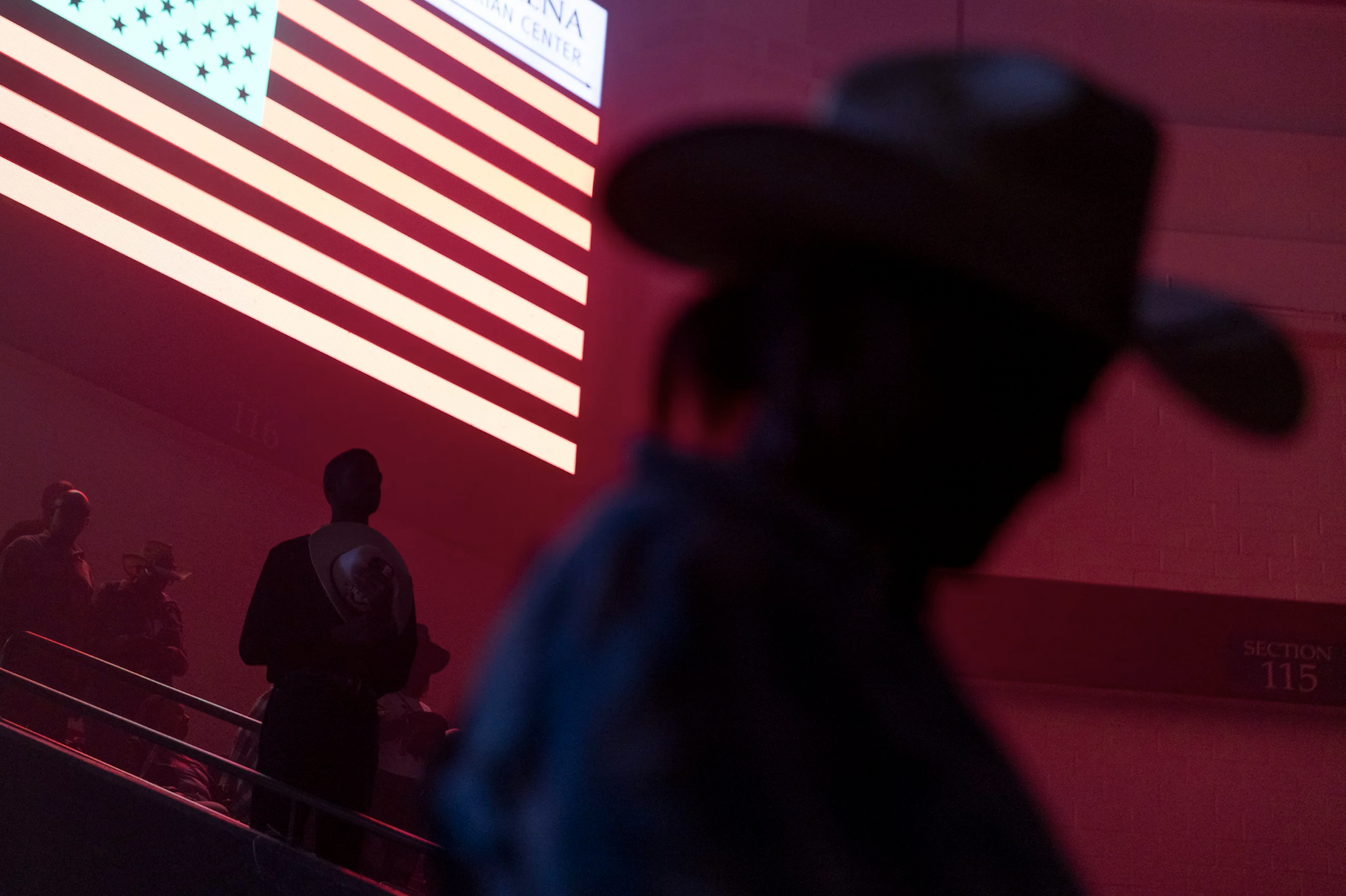 A cowboy hat wearing man, in silhouette stands underneath a big screen displaying the American flag.