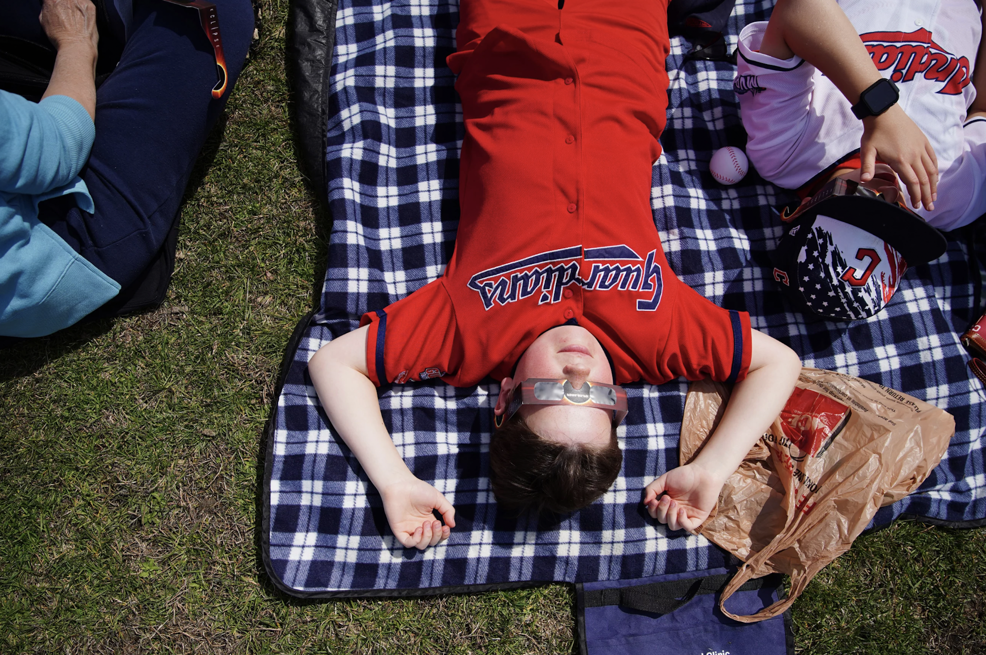 A boy child lying flat on his back on a blanket outdoors, wearing eclipse glasses and a red baseball shirt.