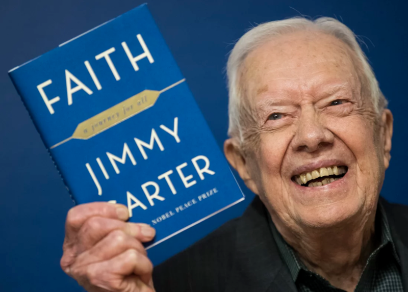 A senior man smiles broadly while holding up a blue book next to his face.