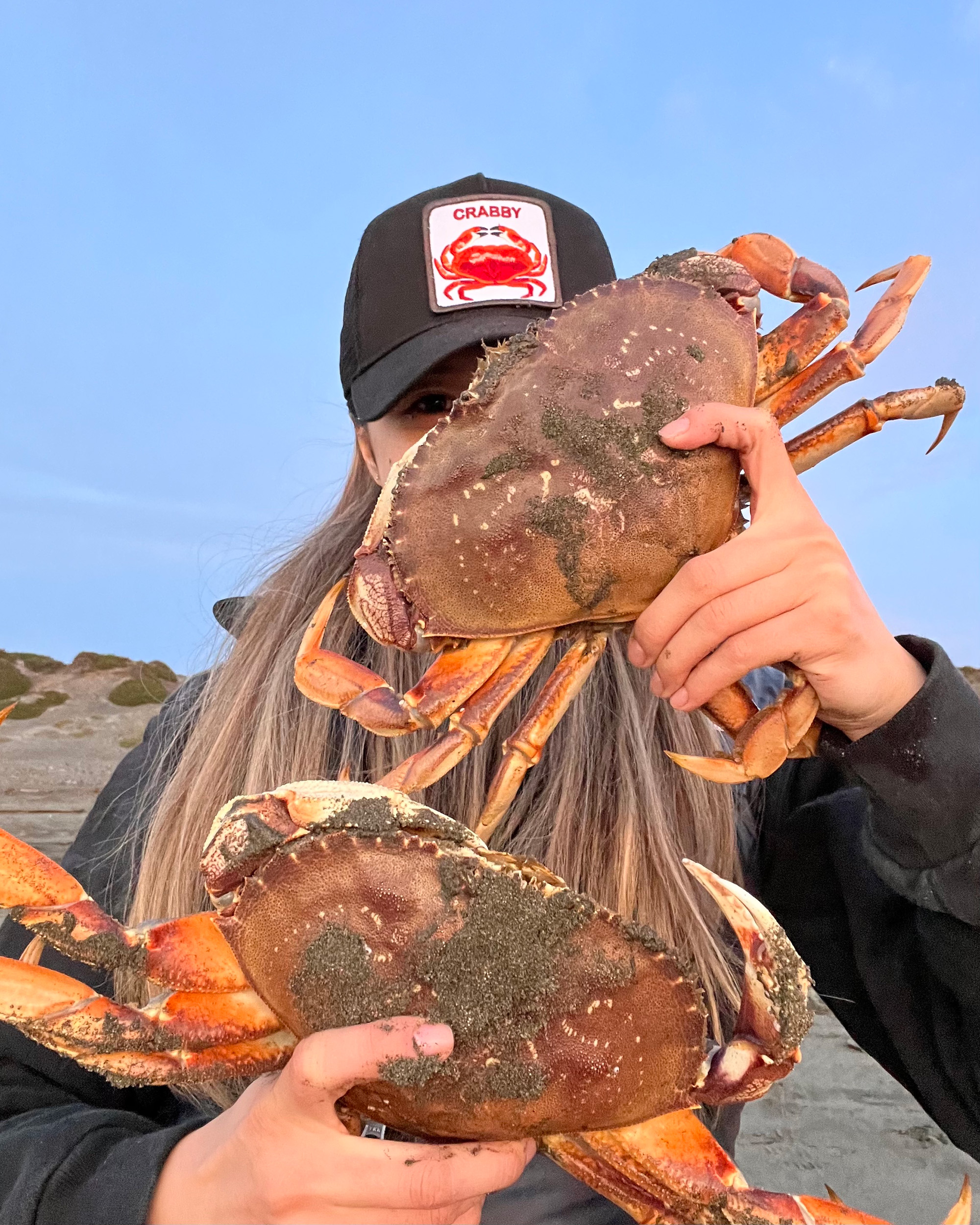 A woman in a trucker hat that reads "Crabby" holds up two large Dungeness crabs.