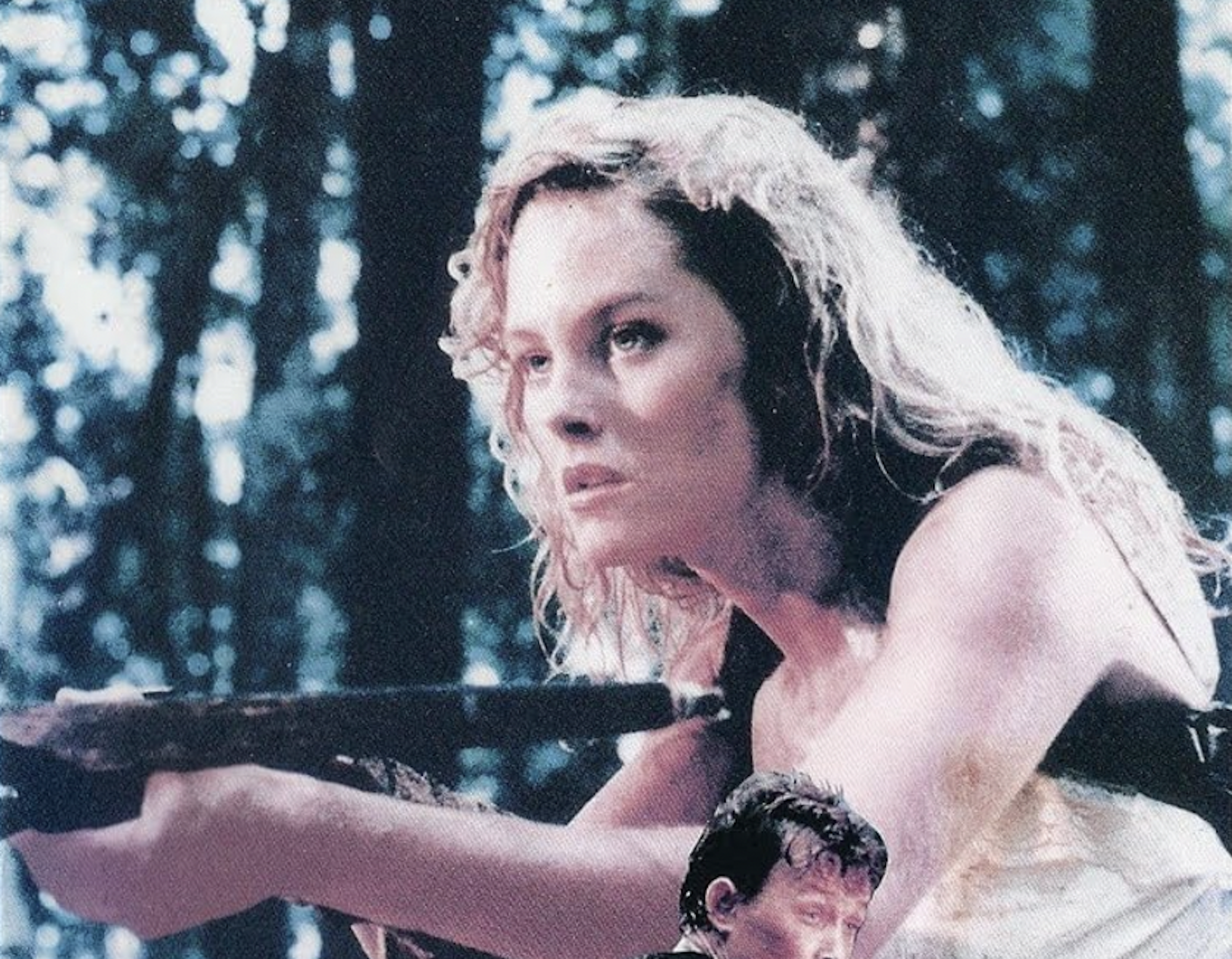 In a forested area, a blond woman holds up a weapon and looks into the distance.
