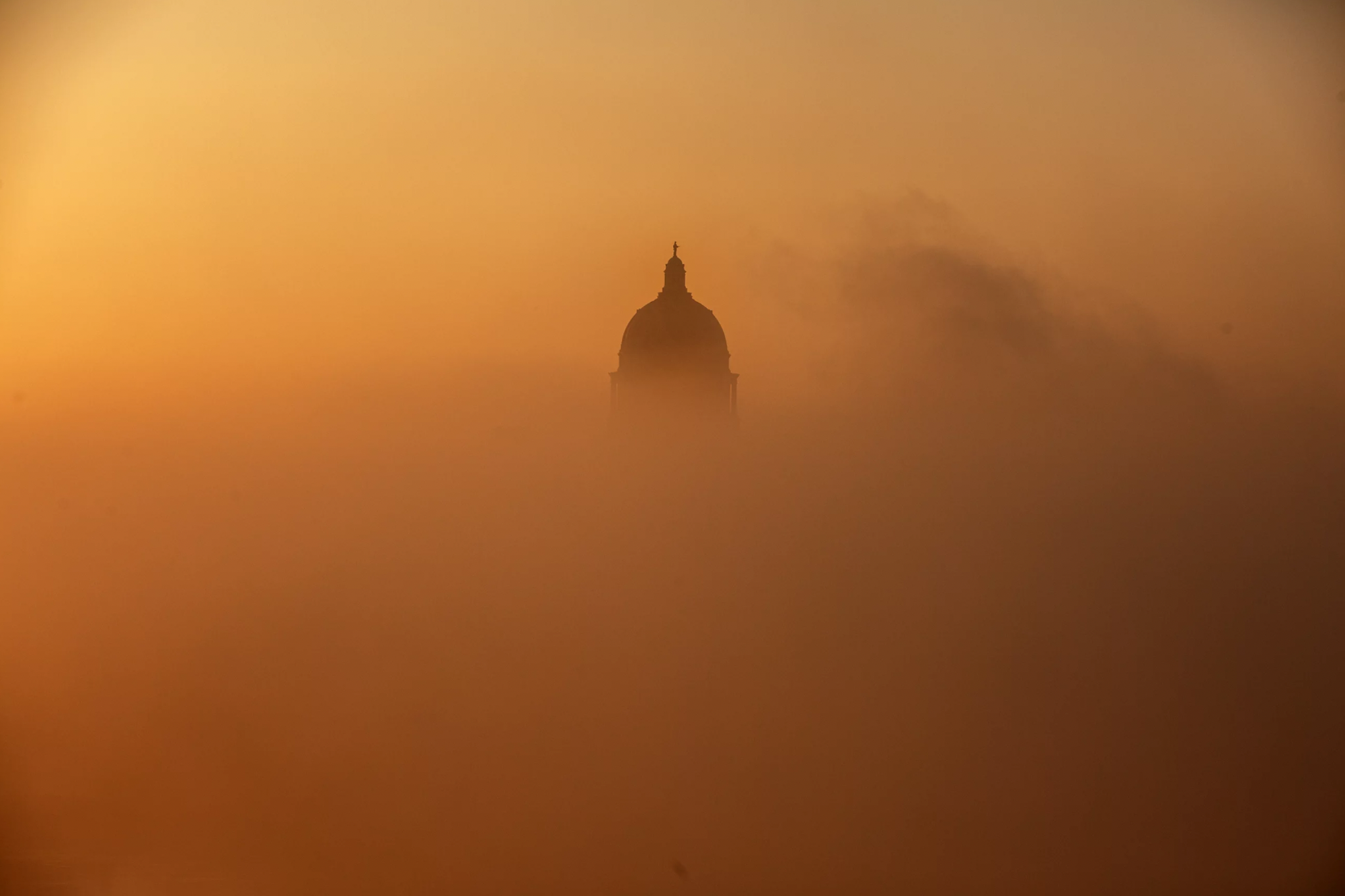 The shape of the top dome of a capitol building rises out of thick orange fog that is obscuring everything else on the ground.