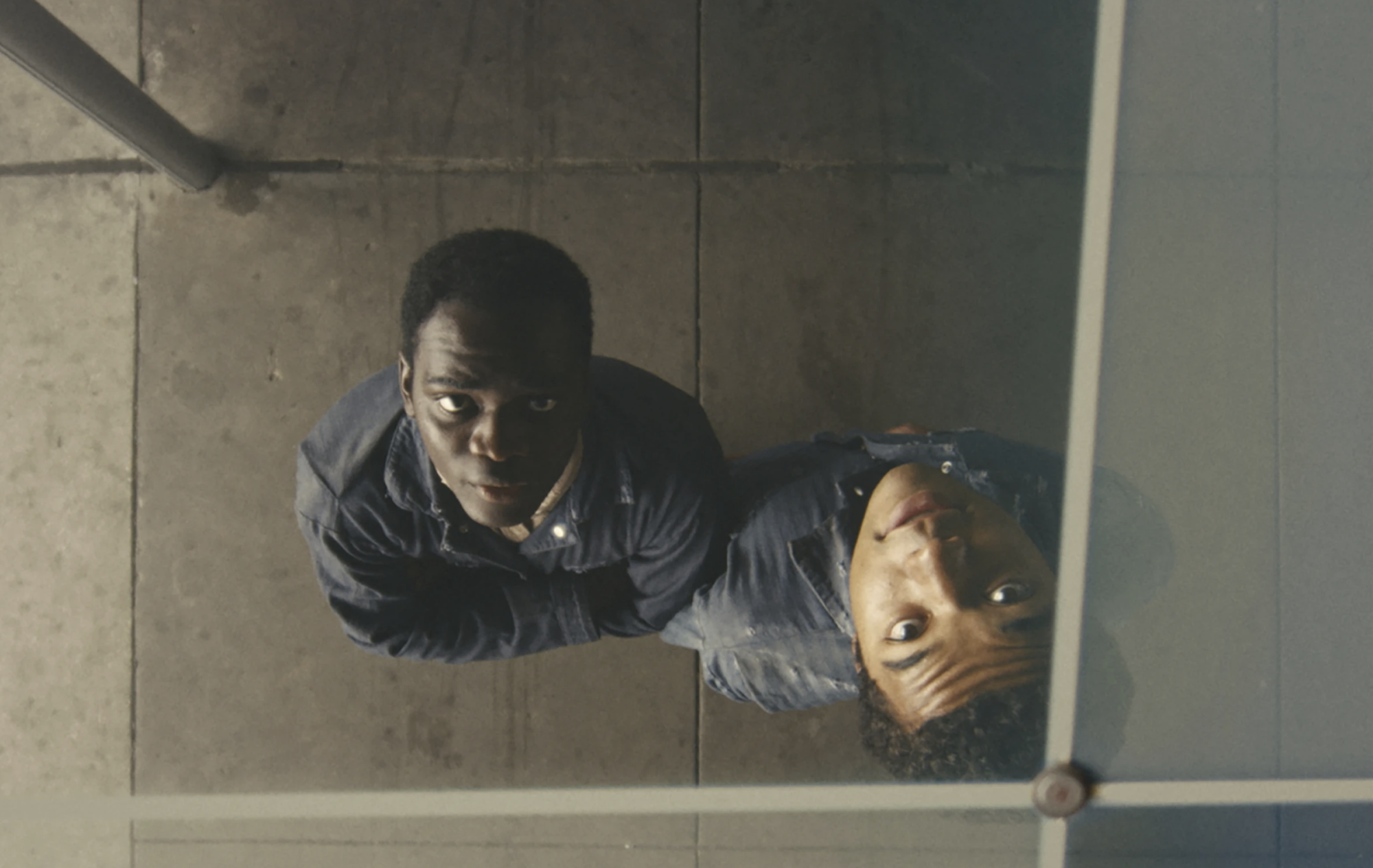 Two young Black men's faces are reflected in a mirror on a ceiling that they are standing beneath.