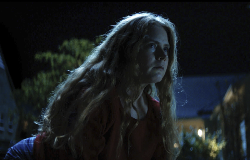 A white woman with long flowing hair crouches down on a lawn under the moonlight.