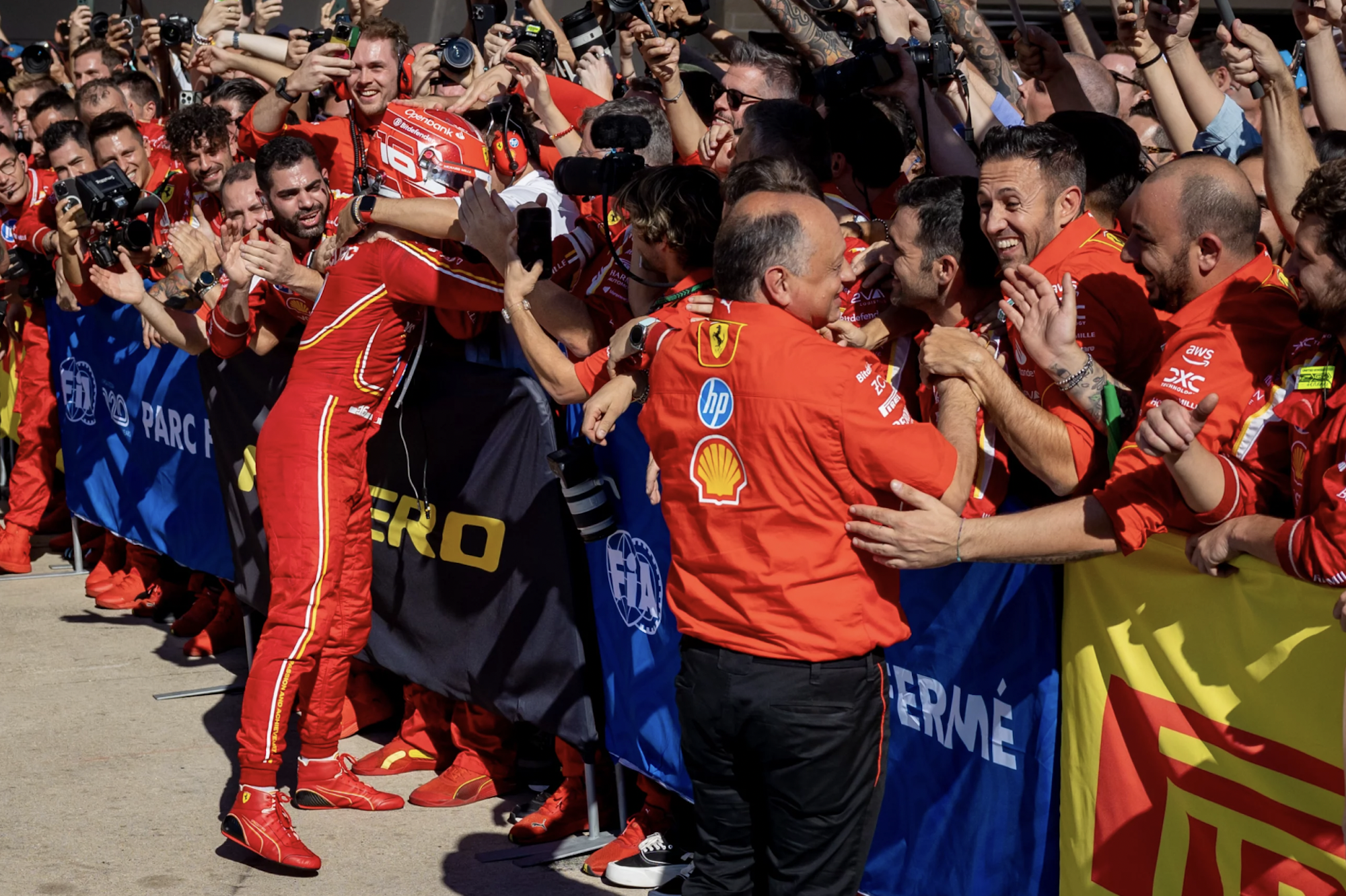 A racing driver in full suit and helmet greets elated fans over a barrier.